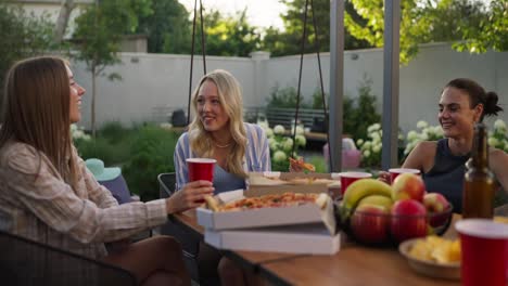 Three-girls-eat-pizza-communicate-laugh-during-lunch-at-a-table-in-the-courtyard-of-a-suburban-house