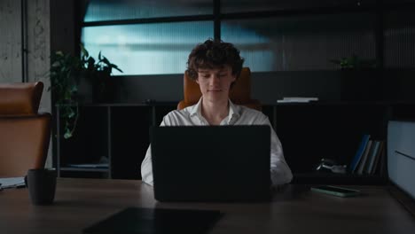 Portrait-confident-young-guy-with-curly-hair-in-a-white-shirt-posing-and-working-at-a-gray-laptop-in-a-modern-office