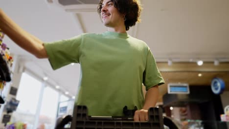 Bottom-view-of-a-happy-brunette-guy-with-curly-hair-in-a-green-T-shirt-puts-necessary-goods-in-a-cart-during-his-shopping-in-a-supermarket