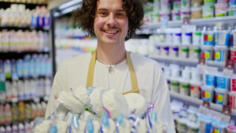 Zoom-Out-portrait-of-a-Happy-guy-with-curly-hair-standing-and-holding-dairy-products-in-his-hands-in-the-department-of-the-supermarket-where-he-works