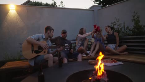 Happy-group-of-five-young-adults-chatting-and-having-fun-during-their-party-near-a-bonfire-in-the-evening-in-the-courtyard-of-a-country-house