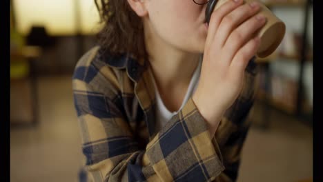 Close-up-of-a-girl-student-with-curly-hair-and-glasses-drinks-coffee-from-a-paper-cup-while-working-in-the-university-library