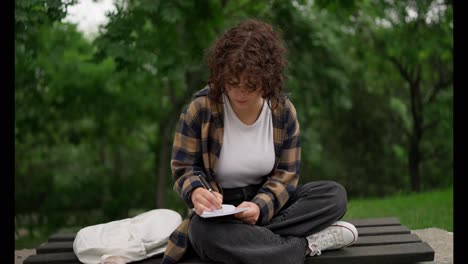 Portrait-of-a-brunette-student-girl-with-curly-hair-in-a-brown-shirt-sitting-on-a-bench-and-making-notes-in-a-notebook-in-the-park