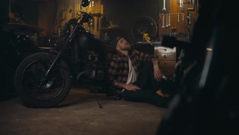 Side-view-of-a-tired-guy-mechanic-in-a-checkered-shirt-wipes-his-forehead-and-leans-on-a-motorcycle-to-rest-in-his-workshop-studio