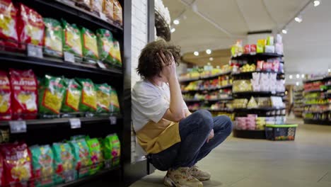 Ein-Müder-Typ-Mit-Lockigem-Haar,-Ein-Supermarktangestellter,-Lehnt-Während-Seiner-Kurzen-Pause-Im-Supermarkt-An-Einer-Wand-Mit-Regalen-Und-Freizeitartikeln