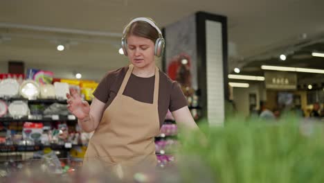 Confident-girl-supermarket-worker-in-a-brown-T-shirt-and-apron-in-wireless-headphones-listening-to-music-and-arranging-goods-on-the-counter-in-a-supermarket