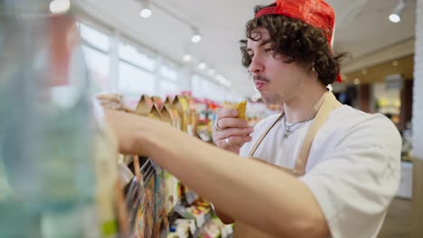 Side-view-of-a-brunette-guy-with-curly-hair-chews-a-bun-and-rearranges-goods-on-the-shelves-during-his-work-in-a-supermarket