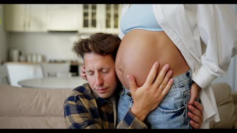 A-middle-aged-man-in-a-plaid-shirt-puts-his-ear-to-the-belly-of-his-pregnant-wife-and-listens-to-a-small-child-during-pregnancy-at-home