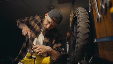 Ein-Fleißiger-Mechaniker-Mit-Bart-Arbeitet-Mit-Einer-Feile-Auf-Einer-Werkbank-Und-Trinkt-Limonade,-Während-Er-In-Seiner-Werkstatt-Ein-Teil-Repariert