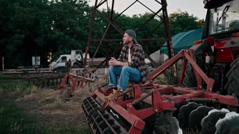 Side-view-of-a-confident-guy-farmer-in-a-cap-holding-a-spikelet-in-his-teeth-and-sitting-on-a-combine-among-greenhouses-on-a-field-on-a-farm