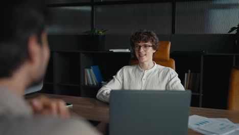 Over-the-shoulder-a-happy-young-guy-with-curly-hair-wearing-glasses-in-a-white-shirt-is-having-an-interview-with-an-experienced-employee-in-the-office