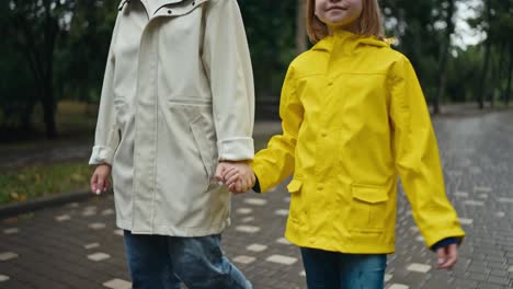 De-Cerca,-Una-Adolescente-Rubia-Con-Una-Chaqueta-Amarilla-Camina-De-La-Mano-Con-Su-Madre-Por-Un-Callejón-En-El-Parque-Después-De-La-Lluvia.