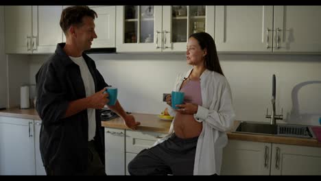 Happy-pregnant-brunette-girl-drinks-tea-and-talks-with-her-husband-during-breakfast-in-the-morning-in-the-kitchen