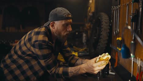 Side-view-of-a-mechanic-guy-in-a-plaid-shirt-drinking-soda-and-eating-a-burger-during-lunch-in-a-studio-workshop