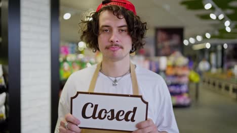 Sad-guy-supermarket-worker-with-curly-hair-holds-in-his-hands-a-sign-with-the-inscription-Closed-while-in-a-closed-supermarket