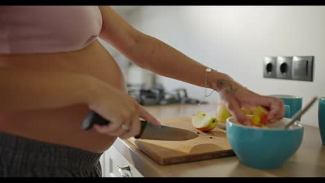 Close-up-of-a-pregnant-woman-cutting-apples-and-placing-them-into-a-bowl-while-preparing-for-breakfast-in-the-morning