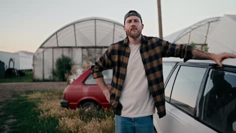 Portrait-of-a-confident-farmer-guy-with-a-beard-in-a-checkered-shirt-posing-near-the-car-among-the-greenhouses-on-the-farm