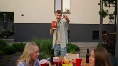 Happy-blond-guy-with-a-mustache-throws-an-American-football-ball-to-his-friend-while-relaxing-at-a-table-in-the-backyard-of-a-house