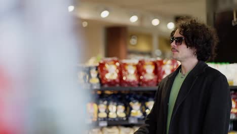 A-confident-and-cool-guy-with-curly-hair-in-black-glasses-and-a-black-coat-chooses-the-products-he-needs-in-a-supermarket,-choosing-from-two