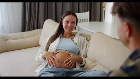 Over-the-shoulder-Happy-pregnant-brunette-woman-communicating-with-her-husband-while-sitting-on-the-sofa-at-home