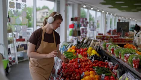 Side-view-of-a-happy-girl-wearing-wireless-headphones-in-a-brown-t-shirt-and-apron-listening-to-music-and-arranging-vegetables-on-the-counter-during-her-work-and-in-the-supermarket