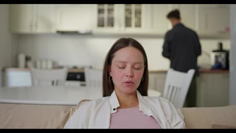 Brunette-pregnant-girl-sits-on-the-sofa-while-her-husband-brings-her-a-cup-of-hot-tea-at-home-during-the-weekend