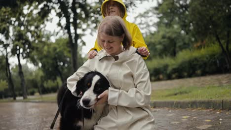 Eine-Glückliche-Blonde-Frau-In-Einer-Weißen-Jacke-Spielt-Mit-Ihrem-Schwarzen-Hund,-Während-Sie-Nach-Dem-Regen-Mit-Ihrer-Tochter-In-Einer-Gelben-Jacke-Durch-Die-Gasse-Im-Park-Spaziert