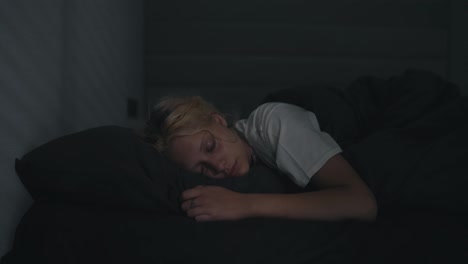 A-young-blonde-girl-in-a-white-T-shirt-sleeps-in-a-gray-bed-in-a-dark-room