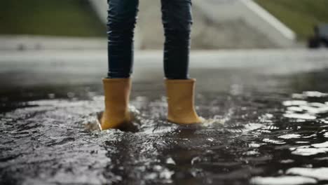 A-teenager-in-orange-boots-actively-walks-through-a-large-puddle-and-pushes-the-water-from-side-to-side-on-the-street-during-the-rain