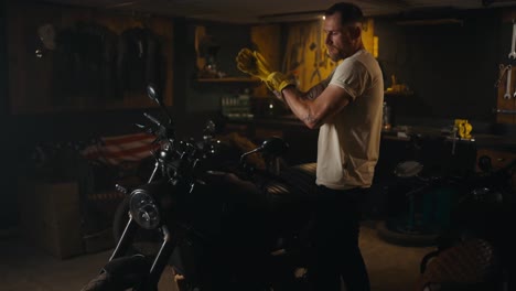 Confident-guy-mechanic-in-a-white-T-shirt-puts-on-yellow-gloves-and-sits-on-his-bike-in-the-workshop-garage