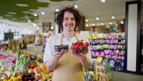 Portrait-of-a-happy-guy-with-curly-hair-a-supermarket-worker-who-holds-in-his-hands-small-boxes-of-blueberries-and-strawberries-in-the-supermarket