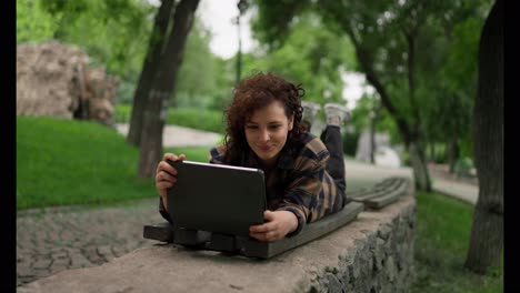 Eine-Glückliche-Studentin-Mit-Lockigem-Haar-Liegt-Auf-Einer-Bank-Und-Schaut-Während-Einer-Unterrichtspause-Im-Park-Auf-Ein-Tablet