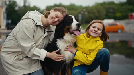 Portrait-of-a-happy-blonde-woman-sitting-with-her-teenage-daughter-petting-their-black-purebred-dog-while-walking-in-the-park-after-the-rain