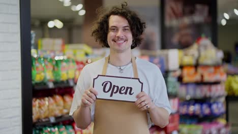 Porträt-Eines-Glücklichen-Kerls-Mit-Schwarzen-Lockigen-Haaren,-Der-Lächelt-Und-In-Einem-Supermarkt-Ein-Schild-Mit-Der-Aufschrift-„offen“-In-Den-Händen-Hält