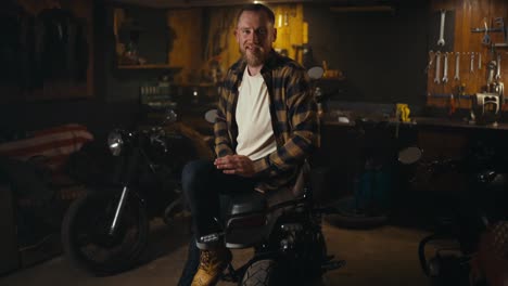 Portrait-of-a-happy-blond-biker-with-a-beard-in-a-checkered-shirt-smiling-and-posing-on-his-bike-in-a-workshop-garage