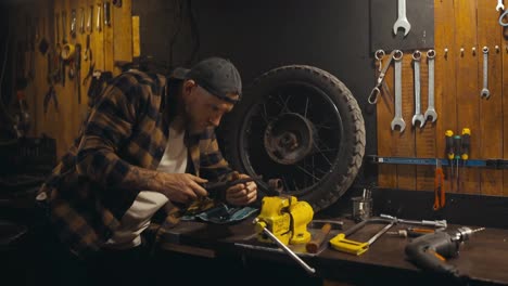 Confident-guy-mechanic-in-a-cap-and-plaid-shirt-works-with-a-file-on-a-clamp-on-a-workbench-in-his-workshop