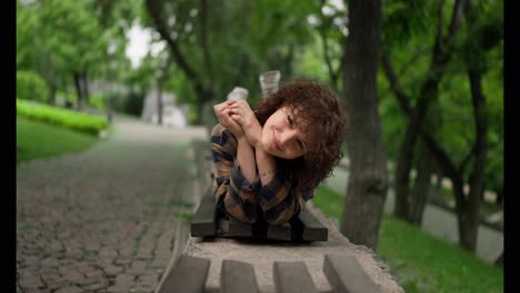 Retrato-De-Una-Estudiante-Feliz-Con-Cabello-Rizado-Acostada-En-Un-Banco-Y-Posando-En-El-Parque.