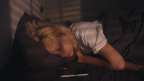 Tired-blonde-girl-in-a-white-T-shirt-sleeps-and-is-woken-up-by-an-alarm-clock-early-in-the-morning-in-a-gray-bed-at-home