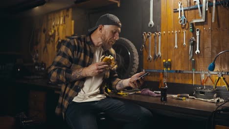 A-blond-guy-with-a-beard-in-a-plaid-shirt-eats-a-burger-watches-a-video-and-drinks-soda-in-his-workshop-studio