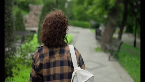 Rear-close-up-view-of-a-girl-with-curly-hair-in-a-plaid-shirt-walking-through-the-park-and-turning-back-during-her-walk-after-her-student-work