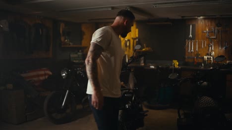 A-blond-guy-in-a-white-T-shirt-inspects-his-motorcycle-in-a-workshop-garage