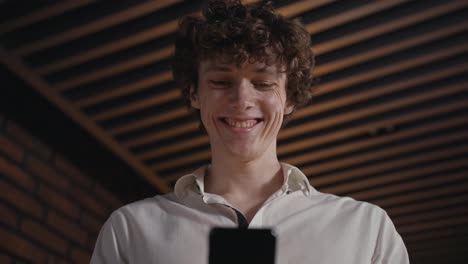 Close-up-of-a-happy-guy-with-curly-hair-in-a-white-shirt-looking-at-the-screen-of-his-smartphone-and-smiling-widely-in-the-office