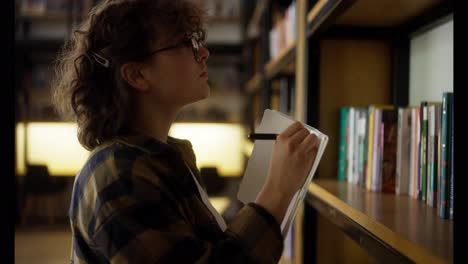 Confident-girl-with-curly-hair-in-glasses-makes-notes-in-a-notebook-using-a-pen-near-shelves-with-books-in-the-library