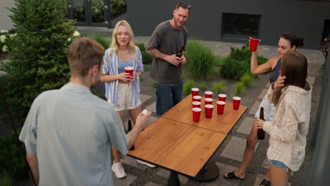 Rear-view-of-a-confident-blond-guy-with-his-company-playing-beer-pong-and-throwing-a-white-ball-on-the-side-during-a-party-in-a-country-house