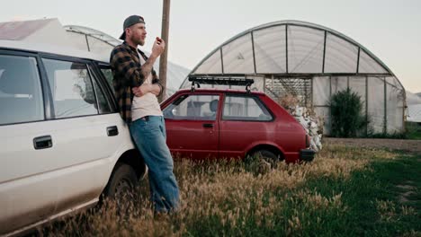 Selbstbewusster-Bauer-Mit-Mütze-Lehnt-An-Einem-Weißen-Auto-Und-Probiert-Die-Tomatenernte-Auf-Einer-Farm-Zwischen-Gewächshäusern