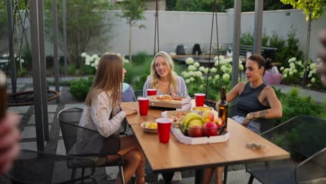 Side-view-of-a-happy-blonde-girl-and-her-two-friends-talking-while-the-guys-clink-glasses-with-brown-bottles-of-beer-and-have-a-good-time-in-the-courtyard-of-a-country-house