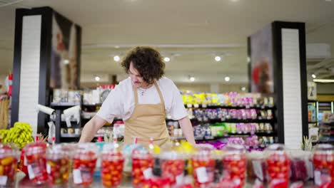 A-confident-guy-with-curly-brunette-hair-in-a-yellow-apron-takes-inventory-of-products-and-rearranges-citrus-fruits-during-his-work-in-a-supermarket