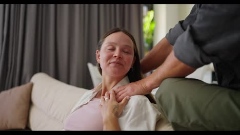 Happy-brunette-woman-sitting-on-the-sofa-while-her-husband-holds-her-shoulders-and-massages-her-at-home-on-the-weekend