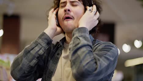 Close-up-of-a-happy-brunette-guy-with-curly-hair-listens-to-music-loudly-on-his-wireless-headphones-and-sings-while-shopping-in-a-supermarket