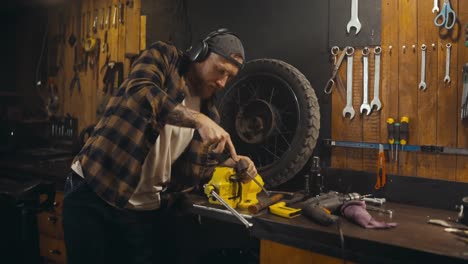 Confident-guy-mechanic-in-headphones-listens-to-music-and-works-while-drinking-soda-from-his-workshop-studio